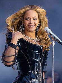 Beyoncé performing on stage, wearing a black haute couture minidress and silver jewelery and looking to her right. In front of her is a silver microphone.