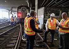 Blick aus dem Gleisbett, ein Dutzend Männer in MTA-Warnwesten arbeiten oder debattieren vor einem abgestellten U-Bahn-Zug.