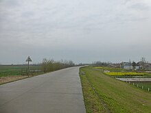 A road over a raised embankment of earth, a marsh on the left side, and some small farms on the other.