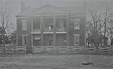 A black and white photograph of the front of an old two story brick building with a large porch and portico behind a wooden fence. A man stands in the doorway, a small painted sign advertising a lawyer's services is hung near the front door, and a gazebo is located to the right of the building.