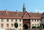 Photographie d'un bâtiment de deux étages avec une arche centrale surmontée d'un balcon en bois, d'un fronton en colombage et d'une flèche en bois