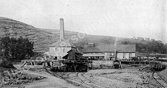 Gravure en noir et blanc : un bâtiments avec toit à quatre pans est doté d'une cheminée de base carré a côté d'un autre bâtiment tout en longueur muni d'un clocheton, des voies ferré passent devant les bâtiments.