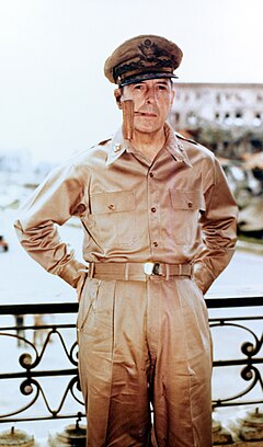 MacArthur in khaki trousers and open necked shirt with five star rank badges on the collar. He is wearing his field marshal's cap and smoking a corncob pipe.