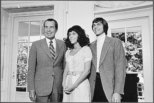 A dark-haired older man in a suit, a young woman with long dark hair wearing a dress and a young man with mid-length hair wearing a jacket over a rollneck sweater