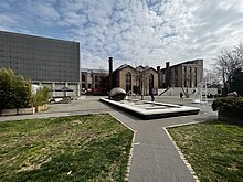 A brick factory stands in front of a park, with open green space, a reflecting pool, and benches