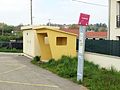 Poteau d'arrêt aux couleurs actuelles du réseau, avec un abribus en béton à côté.