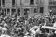 City street with a large group of cyclists facing an open-top car with a man standing inside