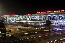 A photo of Hazrat Shahjalal International Airport in Dhaka, Bangladesh at night. The image shows a large airport terminal building illuminated from within. There is a mostly empty parking lot in front of the terminal. Text in Bengali, English, and Arabic identify the building as the Hazrat Shahjalal International Airport. The closest green text reads "Terminal-2", while the farthest reads "Terminal-1"