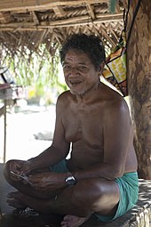 Photo d'un homme assis en tailleur, avec un short vert, le torse nu, sous un abri.