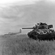 Photo en noir et blanc d'un char d'assaut manoeuvrant dans un champ, avec une carcasse de planeur en arrière-plan.