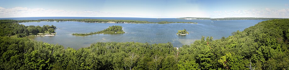 View in August from the now-closed tower. Sawyer Harbor is in the foreground, with the western part of Sturgeon Bay in the background. The small island at right is Heaven On Earth Island, formerly Bug Island. The larger island to the left is Idyll Wood Island. Cabot Point is behind the two islands. It is part of the Idlewild area. In the far distance at center-right is the rock cut of the Old Stone Quarry on the northwest shore of Sturgeon Bay, once the largest in the state.