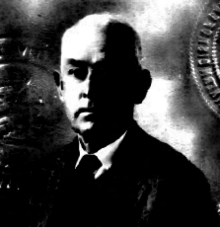 Image: A black-and-white photo of a middle-aged Anglo man with pale receding hair and a somewhat serious expression, wearing a dark tie and sport coat, but no eyeglasses in this picture.