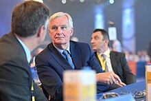 Three men, seated at a conference table in formal suits: Barnier, central, turns to listen to one with his back to the camera.