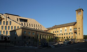 Foto eines Gebäudes mit einem Turm mit Uhr, dahinter ein moderneres Gebäude