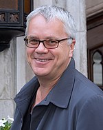 Upper torso of a grey-haired man wearing glasses and a dark grey trench coat.