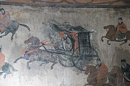 A mural on a wall which shows a man riding a chariot in the center, one person on horseback each is on the charioteer's top right, top left, bottom left; two people on horseback are to his bottom right