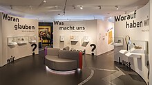 Im Halbkreis angeordnete Ausstellungswände, drei Segmente, die kleine Exponate und je eine Frage enthalten: „Woran glauben wir“ „Was macht uns wütend“ und „Worauf haben wir Lust?“ In der Mitte eine runde Sitzgruppe