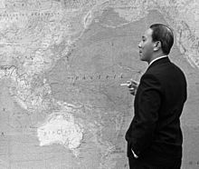 Middle-aged black-haired man, stands side-on in a dark suit with a cigarette in right hand and left hand in pocket, looking at the large map of the Asia Pacific region on the wall.