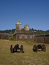 Photograph of the reconstruction of the Fort Ross Chapel, a wooden walled and roofed building with an evergreen-covered hillside behind.