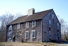 A modern photograph of an old, unadorned two story house clad in weathered shingles