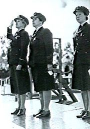 Trois femmes en uniforme militaire sombre, debout au garde-à-vous sur une estrade.