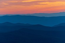 Five mountain ridges in shades of dark blue below an orange and yellow sunset.