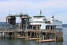 A ferry boat stopped at a wooden pier with a bridge, pilings, and a tower.