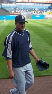 A young man wearing gray baseball pants, a navy-blue pullover windbreaker, and a black baseball glove (on his left hand) steps down from a dirt-and-grass field.