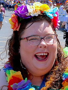 A close up photo of a white woman wearing a multicoloured floral headband, a multicoloured floral garland, and rectangular glasses. She is laughing. She has long hair, and through the hair, you can see a glimpse of her dangly earrings. The photo was taken on a sunny day. There are many people in the background walking, with rainbow coloured flags and banners.