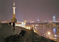 Ludzie spacerujący nocą na placu pod oświetloną kolumną ze statuą Zwycięzcy - nagiego mężczyzny trzymającego miecz. W tle rzeka i panorama miasta.