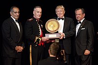 A ceremony in which Trump is receiving the 2015 Marine Corps–Law Enforcement Foundation's annual Commandant's Leadership Award. Four men are standing, all wearing black suits; Trump is second from the right. The two center men (Trump and another man) are holding the award.