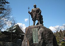 Photo couleur d'une statue en pied d'un homme sur un rocher sur fond de ciel bleu.