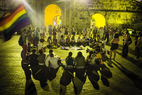 A yellow hued photo of people sitting a circle outside in plaza. A rainbow flag is waving off to the left