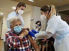 Elderly woman rolls up sleeve as two nurses administer a vaccine.