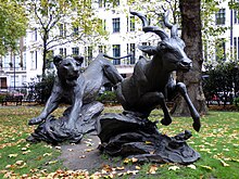 A picture of two dark metal realistic statues. The statue in the foreground shows the front of a leaping kudu and the statue in the background shows the full body of a leaping lioness chasing the kudu. The statues stand in a grass patch of a park in a city.