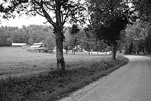 A black and white photograph of a small farm, including a barn, outbuildings, and pasture, taken from a gravel road.