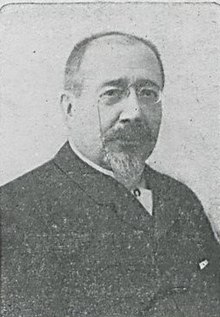 Portrait photographique d'un homme portant une barbiche et de fines lunettes.