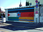 Seitliche Farbfotografie von der Berliner Mauer mit einem Bild, das aus drei Flaggen besteht. Links die palästinensische Flagge und rechts die deutsche Flagge und quer über beide befinden sich zwei blaue Linien und ein blauer Davidstern der israelischen Flagge. An den Seiten der Flagge stehen lange Texte und über dem Bild steht „März 90 Vaterland“. Links neben der Mauer ist ein Durchgang und im Hintergrund sind die Oberbaumbrücke und ein Haus zu sehen.