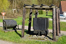 Une berline fleurie posée sur un tas de schistes sous des boisages portant une lampe et les symboles de la mine ; à côté une berline basculante elle aussi garnie de fleurs et entourée de rondins de bois.