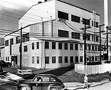 A large four-storey building. The chimney is in the background. There are power poles and power lines in front.