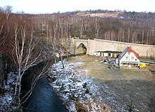 Barevná fotografie s pohledem na koryto řeky s kamenným mostem, domkem a břízami