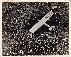 Black-and-white photograph of an enormous crowd of people, packed like sardines, crowding around a small aircraft marked NX211 on the starboard wing