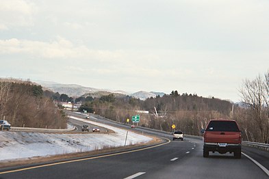 A four-lane highway in snowy weather, curving left with several cars on it. An exit sign and mountains are in the distance.