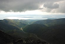 Pohled do zelenošedého údolí s prudkými srázy, vzadu rozsáhlá vodní plocha