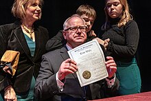 Tim Walz sitting and holding a document. Gwen, Tim, and Hope Walz stand behind him.