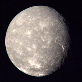 A round spherical body is almost fully illuminated. The surface has a mottled appearance with bright patches among relatively dark terrain. The terminator is located near the right edge. A large crater can be seen at the terminator in the upper half of the image. Another bright crater can be seen at the bottom. A large canyon runs from the darkness at the lower-right side to visible center of the body.