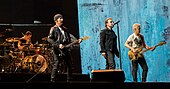 Four men performing on a stage in front of a crowd; two are standing at the front of the stage holding guitars, one in the center is holding a microphone, and one is sitting behind a drum set.