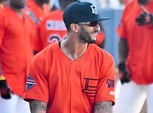 Portrait d'un homme habillé d'un maillot de baseball orange, de lunettes de soleil et d'une casquette noire.