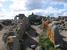 intérieur de l'allée couverte, en pente, vue depuis son entrée. La plupart des dalles de couvertures manquent. En arrière-plan, au sommet de l'éminence, on aperçoit le dolmen.