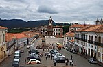 Two-storied white buildings with red roof tiles lining a square. At the end of the square there is a prominent building with a clock tower.